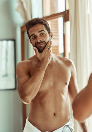 Man, bathroom and self care in mirror in morning for grooming routine, cosmetics and skincare for beard. Person, reflection and cleaning face for glowing skin, hygiene and happy with facial treatmentの写真素材