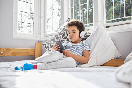Bed, phone and boy with headphones for listening, hearing and ebook for learning with children stories. Home, bedroom and kid with mobile app for games, cartoon and streaming videos for curiousの写真素材