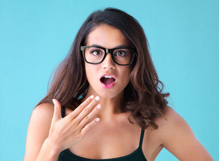 Shocked woman, portrait and surprise with glasses for news or gossip in studio on a blue background. Young female person or model in wow, gasp or omg with facial expression or spectacles for alertの写真素材