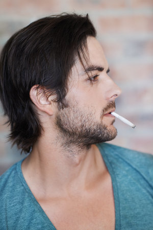 Man, cigarette and urban fashion by brick wall in city for style, edgy and thinking outdoors. Smoking, male person and streetwear by building for addiction, stress relief and relaxation outsideの写真素材