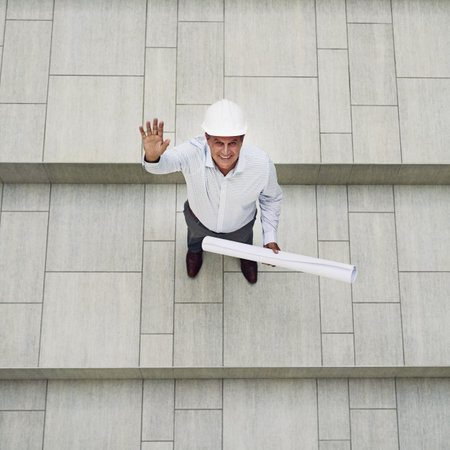 Blueprint, engineering and portrait of construction worker on building site waving from above. Smile, greeting and male industry foreman with architecture plan for renovation, repairs or development.の写真素材