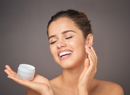 Cream, beauty and face of happy woman for skincare, wellness and facial treatment on gray background. Dermatology, salon and person with moisturizer jar, lotion and anti aging cosmetics in studioの写真素材