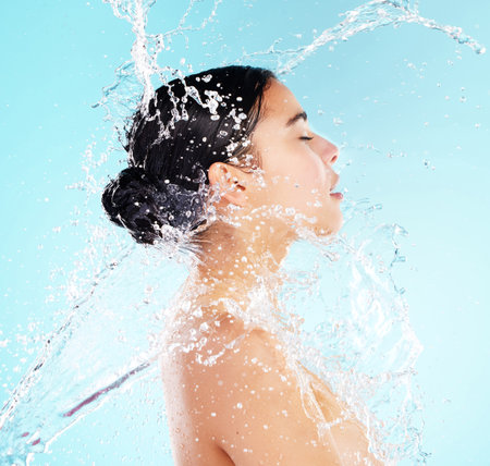 Woman, water splash and profile in studio for beauty with drops, wellness and cleaning by blue background. Girl, model and routine for transformation, grooming and cosmetics with process for skincareの写真素材