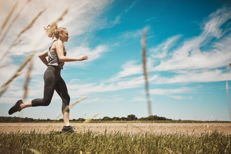 Earphones, fitness and woman running outdoor for healthy body, wellness or exercise to workout. Music, cardio and trail runner training in nature for sport, marathon and jog at park on path in summerの写真素材