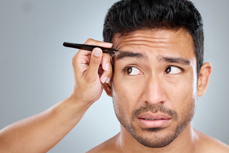 Man, hand and tweezers by eyebrow in studio for grooming, facial epilation and maintenance or skincare. Male person, cosmetic tool and product for hair removal, gray background and hygiene routine.の写真素材