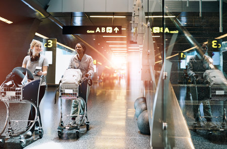 Travel, friends and women at airport with luggage trolley for vacation, holiday or trip together. Walking, suitcase or happy girls at airline with passport for journey, talk or arrival at flight gateの写真素材