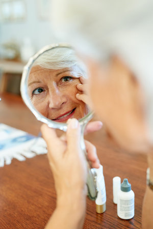 Home, mirror and elderly woman for skincare, checking and face inspection with hand gesture. Reflection, mature person and beauty treatment for wrinkles, anti aging and skin texture in houseの写真素材