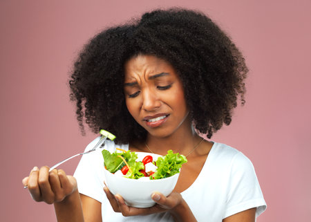 Unhappy, salad and dislike of black woman in studio eating vegetables for nutrition or vegan diet. Girl, pink background and health for food, detox and wellness benefits for weight loss and disgustの写真素材