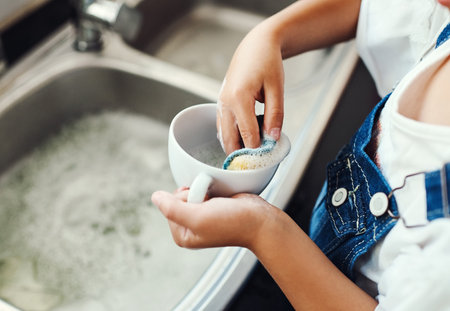 Girl, hands and learning to wash dishes in kitchen, sanitary and prevent bacteria or germs. Female person, child and soap for cleaning mess in home, housework and kid helping for responsibilityの写真素材