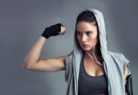 Power, hoodie and woman in studio, fitness and strength of arm with bodybuilding in gym and earphones. Grey background, serious and athlete with music for workout, flexing and sports in health clubの写真素材