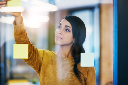 Woman, sticky note and glass wall in office for ideas, planning or brainstorming at company. Person, thinking and paper on board for problem solving, solution or process for job at creative agencyの写真素材