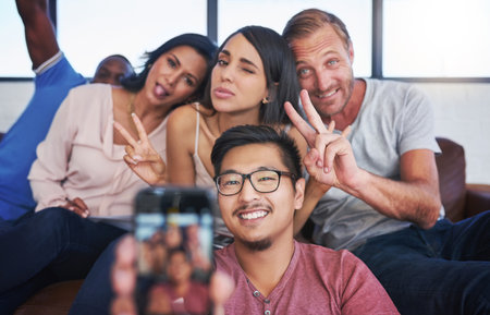 Business people, happy and selfie in office with peace, diversity and sofa for playful, joy and fun. Creative agency, smile and team in workplace with memory for collaboration, cooperation or startupの写真素材