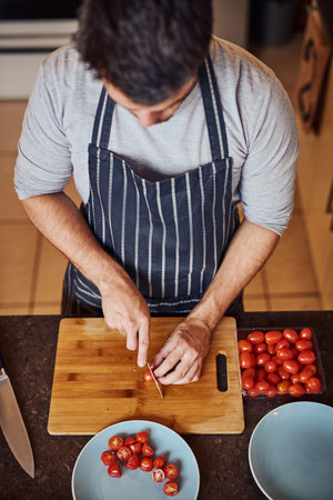 Above, man and cutting with tomatoes for health, nutrition or vegan diet for wellness. Male person, chef and wood board in kitchen for cooking with food and ingredients to prepare home dinnerの写真素材