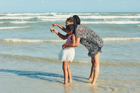 Beach, African mother and child point with blue sky, family and love with happiness and vacation in Miami by ocean. Nature, woman and kid by sea in summer to relax and adventure together with smileの写真素材