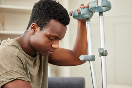 Man, injury and depression on sofa with crutches for healing, recovery and mobility in home. African...