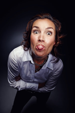 Mocking, tongue out and face of woman with gesture isolated on dark background in studio. Rude, mean...