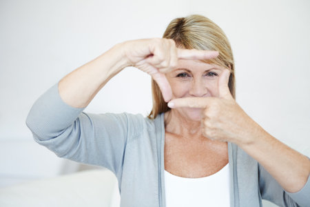 Mature woman, portrait and fingers with frame for picture, moment or capture on sofa at home. Senior female person with gesture, eyes or face for photography, memory or sight in living room at houseの写真素材