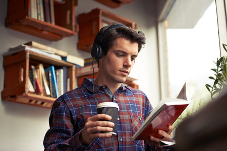 Man, headphones and tea for book at cafe, literature and student to study on campus. Male person, reading novel and learning english for journalism at coffee house, espresso and podcast for fictionの写真素材