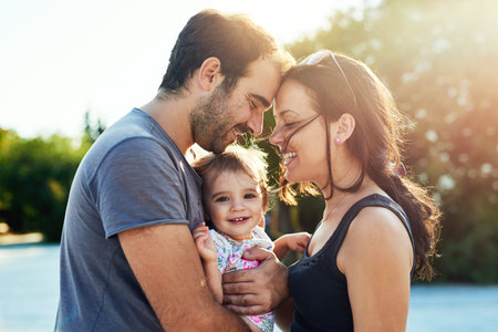 Happy, love and parents with baby in park for bonding, loving relationship and relax together outdoors. Family, sunshine and mom, dad and child for embrace, affection and forehead touch on weekendの写真素材