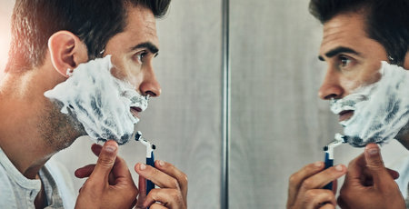 Man, mirror and foam for shaving in bathroom, skincare and hair removal for cleaning in home. Male person, reflection and soap or cream for grooming beard, cosmetics and blade for facial treatmentの写真素材