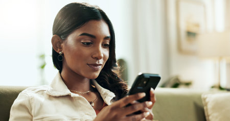Phone, relax and text message with Indian woman on sofa in living room of home for internet search. App, social media and wellness with person typing on mobile in apartment for weekend communicationの写真素材