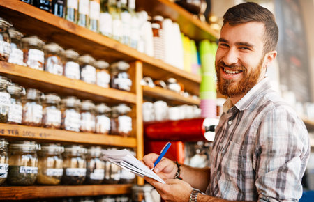 Man, writing and shop owner in portrait at cafe by shelf, paperwork and inspection for stock with smile. Person, entrepreneur and documents with notes, inventory and restaurant management in Turkeyの写真素材