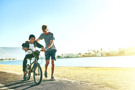 Outdoor, ride and siblings with bicycle for learning, paddle or coordination with balance by space. Lagoon, children or boys with bike for speed, moving or teenage brother with assistance for supportの写真素材
