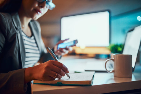 Business, woman and writing in notebook in office at night for planning schedule, project deadline or calendar. Professional, employee or journal for agenda, notes or overtime at desk with smartphoneの写真素材