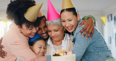 Family, hug and celebration with grandmother on birthday for surprise, present and special event at home. Party hat, happy people and senior woman with gesture of love for cake, candles and dessertの写真素材