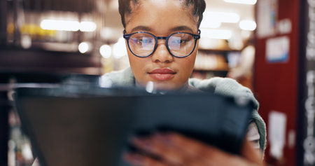 African, girl and student with tablet in library for research information, university news and study schedule. Woman, digital and reading glasses with education ebook, exam results and project reportの写真素材