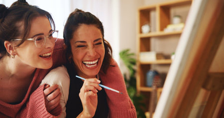 Lesbian, couple and laughing with canvas in home for painting project, help and hobby on weekend. Happy, LGBTQ and women with hug by easel in house for love, support or discussion on creative artworkの写真素材