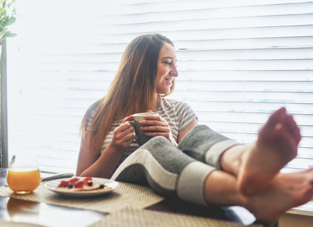 Drinking coffee, relax and woman with feet on table, idea or thinking of eating healthy breakfast. Tea, smile or girl with legs up in home for peace, calm or enjoy morning espresso beverage by windowの写真素材