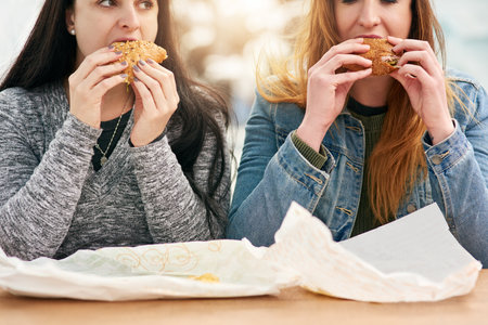 Women, together and eating sandwich in city, park and share with snack, hungry or diet in summer. People, bread and relax with fast food, sub and friends in sunshine with reunion for bonding in Spainの写真素材