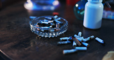 Drugs, addiction and ashtray on table in dark apartment with opioid crisis, crime and danger. Risk, substance abuse and cigarette with container of pills, narcotics or toxic habit in home at nightの写真素材
