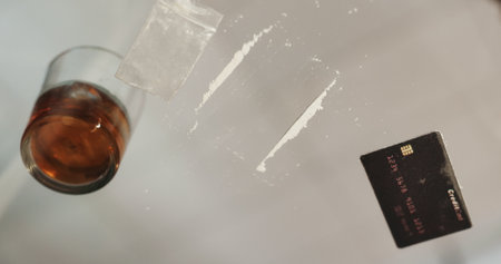 Drugs, cocaine and alcohol on glass table in apartment with opioid crisis, crime and narcotic bag. Risk, substance abuse and credit card with drink, white powder lines or toxic habit in home at nightの写真素材