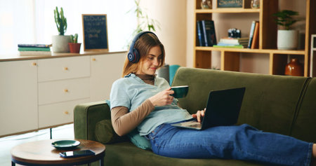 Laptop, headphones and girl on sofa with coffee, comfort or listening to online webinar in home....