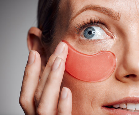 Closeup, woman and eye patches for skincare, beauty and collagen treatment. Apply mask, hand and mature model with cosmetics on face for dermatology, benefits and anti aging on gray studio backgroundの写真素材