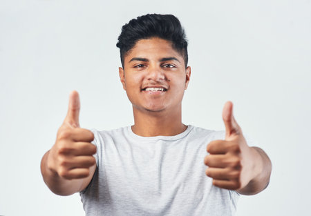 Indian, portrait and happy man with thumbs up for winning, good job or well done on a white studio background. Young, male person or gen z model with smile, like emoji or yes sign for OK or greatの写真素材