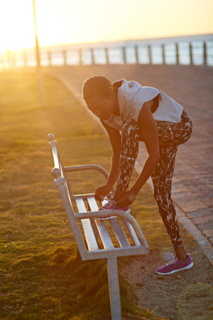 Black woman, runner and tie shoes on park bench by sea with sunrise, sport and promenade workout....
