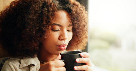 Woman, morning and blowing coffee by window in home, relax and memory with drink at apartment. Person, reflection and thinking with cup for tea, latte and perspective with caffeine beverage in houseの写真素材