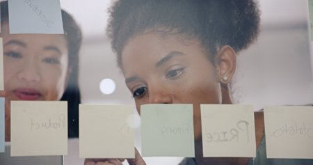 Business, women and sticky notes on glass wall for planning, strategy or brainstorming. People, employees and ideas as partners for teamwork, collaboration or coworking for project deadline at officeの写真素材