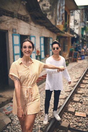 Couple, portrait and smile with walking on train tracks in Vietnam for travel adventure, local experience or love. Tourist attraction, interracial people and holding hands on railroad for sightseeingの写真素材