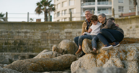 Grandparents, child and happy on rocks at beach with hug, memory and outdoor on holiday with bonding. Senior man, woman and boy with embrace, care and love with family on vacation with view in Italyの写真素材