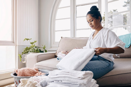 Fold, cleaning and woman in home for laundry for organizing clothes and washing in living room. Cotton, basket and housekeeping cleaner with fabric, garment or towel in morning routine for wellnessの写真素材