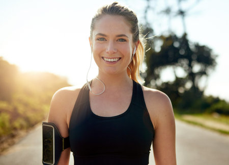 Happy woman, portrait and runner with earphones on road for workout, cardio or outdoor exercise. Young, female person or athlete with smile for music, podcast or fitness on asphalt, path or streetの写真素材