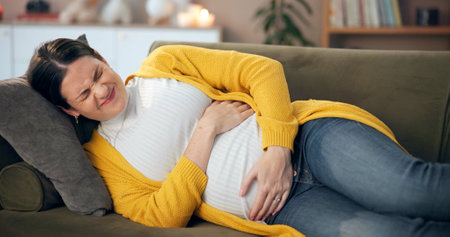 Pregnant, woman or stress in home with pain, cramping or hold stomach for relief on sofa. Future mom, rest or discomfort in living room for contractions, bloating or prenatal care with worry for babyの写真素材