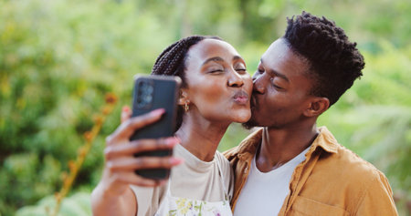 Black couple, outdoor and selfie with kiss in park with holiday, love and care together with social media and smile. Happy, people and bonding on summer vacation with profile picture in garden on webの写真素材