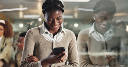 Black woman, phone and texting by window, office or chat at night with reflection, contact or networking at company. African person, laugh and smartphone on app, happy or reading at creative agencyの写真素材