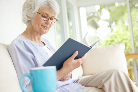 Elderly woman, happiness and reading book for education in retirement home on weekend on break. Senior, female person and glasses as confident, cheerful and calm in living room on couch for knowledgeの写真素材