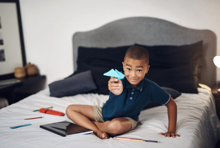 Boy, portrait and bed with paper plane for fun creation, activity or creativity at home. Young, child or kid with smile or playing for flight, airplane or invention for childhood in bedroom at houseの写真素材